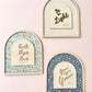 Three arched decorative wall art pieces mounted on a pale pink wall. Each piece has a light wood frame and a central cream panel with inspirational text: one says 'Be the Light,' one says 'Faith Hope Love,' and one says 'Grow upon Grace.