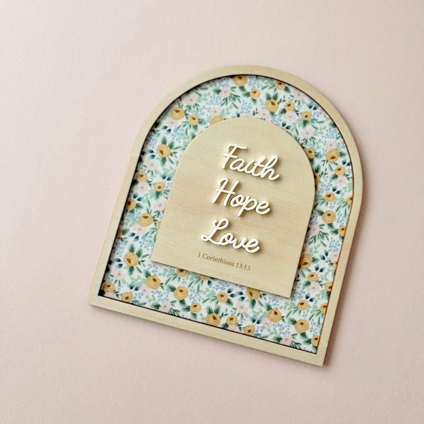 An arched wooden plaque with a floral patterned background and a central wooden tab labeled 'Faith Hope Love' with '1 Corinthians 16:14' underneath, displayed on a light beige wall.