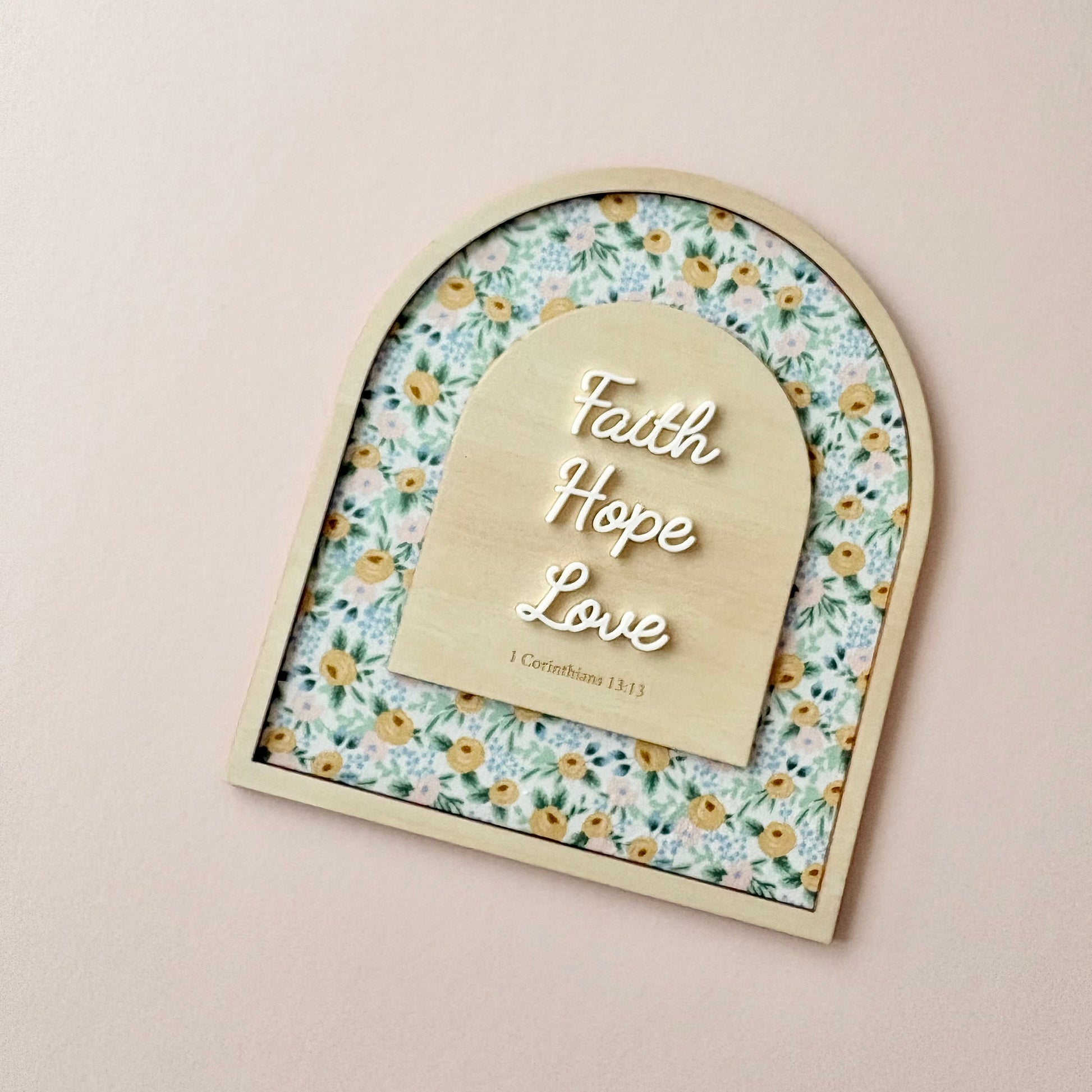An arched wooden plaque with a floral patterned background and a central wooden tab labeled 'Faith Hope Love' with '1 Corinthians 16:14' underneath, displayed on a light beige wall.