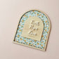 An arched wooden plaque with a floral patterned background and a central wooden tab labeled 'Faith Hope Love' with '1 Corinthians 16:14' underneath, displayed on a light beige wall.