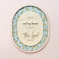 An oval wooden wall plaque featuring a floral patterned border and a central light wood panel with the text 'As for me and my house we will serve the Lord' and 'John 14:23' in a clean, modern font, displayed against a soft pink background.